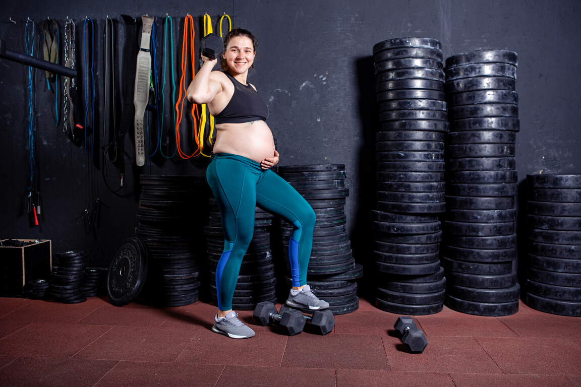 Active sports pregnancy. Workout in the gym together during pregnancy. Sports and a healthy lifestyle during pregnancy. Pregnant professional female athlete posing with fitness equipment background